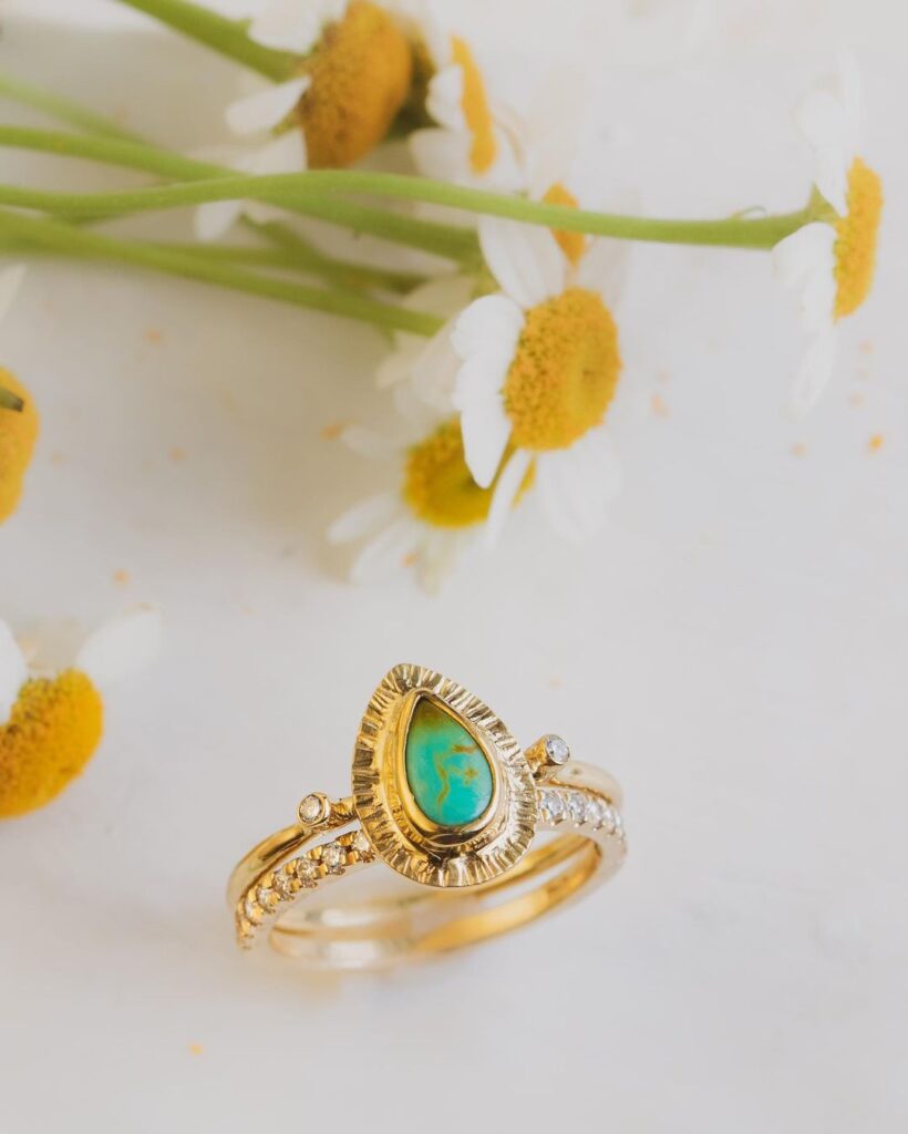 a set of gold bands with small diamonds and a turquoise tear-drop center stone next to some daisies