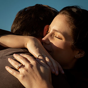 A couple embraces with De Beers Desert Diamond engagement ring visible on one person's hand.
